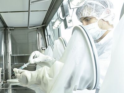 Die Innenkammer eines Isolators aus der seitlichen Perspektive. Die Rückwand ist geschlossen. An der rechten Seite sitzt außen eine Person in Schutzkleidung, die durch Handschuhe nach innen greift. Dort hält die Person eine Spritze.