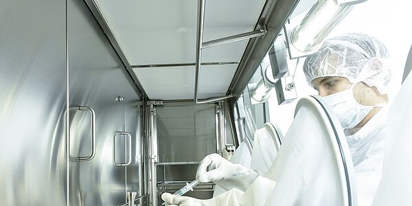 Die Innenkammer eines Isolators aus der seitlichen Perspektive. Die Rückwand ist geschlossen. An der rechten Seite sitzt außen eine Person in Schutzkleidung, die durch Handschuhe nach innen greift. Dort hält die Person eine Spritze.
