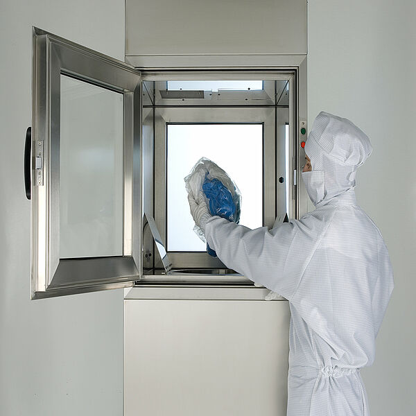 A person in white protective clothing including protective hood, mouth guard and protective gloves is standing in front of a square door with a glass pane that is open. The door is exactly on her outstretched hand. In the door frame are a green and a red button. In her left hand she holds a transparent plastic bag with white and blue crumpled cloth and protective gloves.