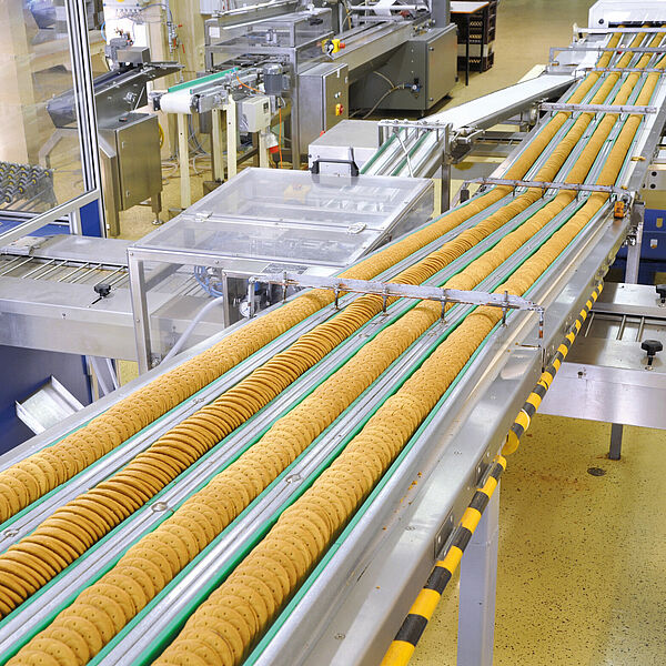 On a conveyor belt running from the lower left corner to the upper right corner, four rows of cookies are stacked one after the other. To the left of the conveyor belt is another small one. Another conveyor belt also passes horizontally under the first run.
