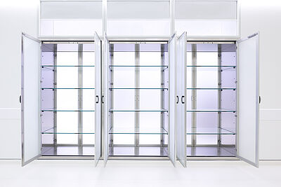 Drei in eine Wand eingebaute sehr hohe Schränke. Jeder Schrank hat zwei Türen mit schwarzen Griffen, die geöffnet sind. In jedem Schrank sind vier Regalböden aus Glas.
