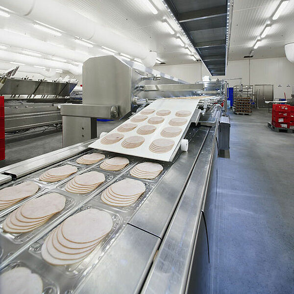  There are three rows of plastic packaging on an assembly line. Inside these plastic packages are sliced sheets of sausage. Approximately in the middle of the conveyor belt there is a beveled plate on which a white conveyor belt is mounted. On it lie cut portioned sausage leaves. Above the conveyor belt, hanging from the ceiling, runs a narrow box, on the underside of which is attached a fine grid, separated by several plates.