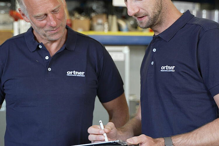 Zwei Männer in Ortner Polo Shirts stehen nebeneinander und schauen auf ein iPad, das einer der beiden mit einem Stift bedient.