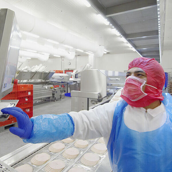  On the right of the picture is a man wearing white protective clothing. Over this he wears a blue apron, blue protective gloves, a red protective mask and a red protective hood. His right hand rests on a plate on which a screen is mounted. In the background, three rows of plastic packaging are lined up on an assembly line. Inside these plastic packages are sliced sheets of sausage. Above the assembly line, hanging from the ceiling, runs a narrow box with a fine grid attached to its underside, separated by several panels. In the man's background, at the end of the room, is a cart, with ten shelves. In six of these shelves lie sausage sticks.