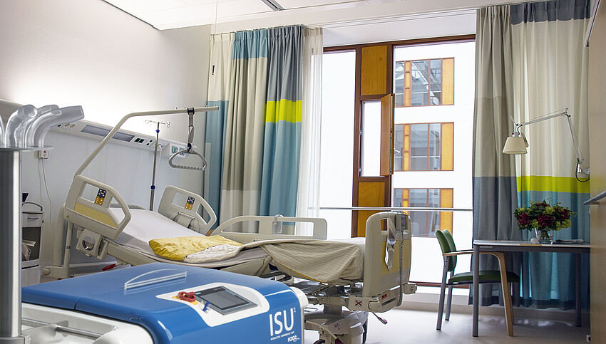 In einem Zimmer steht links ein Krankenbett und rechts ein Tisch, auf dem Blumen stehen. Links vorne steht eine ISU.