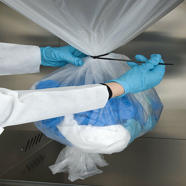 A transparent plastic bag hangs from the top, knotted at the end. Inside is white cloth and blue protective gloves. From the left, there are two hands wearing gloves. The content of the bag above is tied with a black cable tie. 