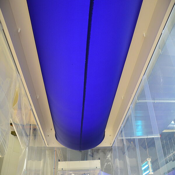  On one ceiling is a white rectangular bar. Between the two longer strips hangs a thick hose, which is made of blue fabric. You can see the underside of the hose. On the left and right, as well as at the bottom of the picture, there are transparent, smooth curtains.