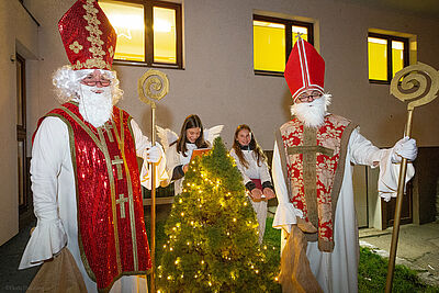 Two Saint Nicholas are standing next to a small Christmas tree. In the background are two girls dressed as angels.