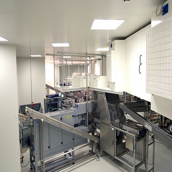  In the center of a large white room are boxes that are connected to each other. From right to left, conveyor belts run upwards. Above, panels hang from the ceiling, on which white cuboid boxes are built. In the right half of the picture, a white box is placed in the foreground; on the front is a rough grid from which a white fabric shows through. A bit further back, the same box is attached.