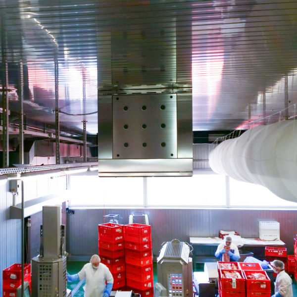  In the lower part of the picture, several stacked crates are distributed around the room. Between them are three people wearing white coats, white face masks and white protective hoods, and blue protective gloves. One of them is standing at a gray tall machine that goes up in a cylinder shape. The lower part is slightly wider and has holes. The second person stands behind the crates and has a large piece of meat in both hands. The third person is walking from left to right. In the middle of the picture, a white wide hose runs along the ceiling to the back. In the center of the ceiling hangs a silver cube. The top edge of the cube is mounted directly to the ceiling. The left, right and bottom edges of the cube are wide strips, so that the central part forms a square area. There are three rows with three holes each.