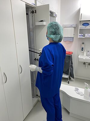 A person in a blue protective suit with white gloves and a green protective hood is standing in front of a cabinet. She is opening one of the narrow doors.