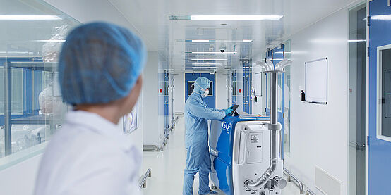  A large bright hallway. On the left is the torso of a person dressed in white with protective hood. In the background, a man in a blue protective suit, protective hood, blue gloves and white mouth guard is standing at a white-blue box on rollers. The box goes up to the person's chest. On the front of the box you can see ISU. Above the box is a tablet on which the person is working. On the side of the box, at the bottom, there is a tube that protrudes up along the box. The tube is about one and a half times as long as the box is high. At the end of the tube is a plate where six smaller curved tubes protrude.