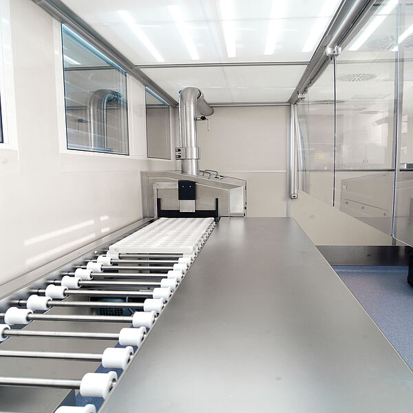  In the left wall, two glass panes are installed at the back towards the corner. Along the wall is a silver long table divided into a closed and an open area. The open area is a conveyor belt made of silver rods with white rollers at the ends. At the last piece towards the end of the room, there is a white flat plate. Just before the end of the room, there is a box that reaches about halfway up the room. The conveyor belt leads into this box through the door that is open at the top. Above the box, a thick silver pipe leads upward. It makes a bend just before the ceiling and eventually leads into the wall. On the right side of the table is a glass wall hanging from above, reaching to about the middle of the height of the room. Translated with www.DeepL.com/Translator (free version)