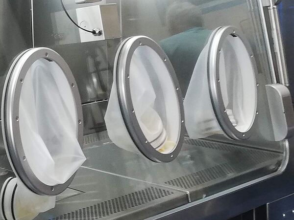 In der Frontscheibe eines Isolators sind vier Handschuhe angebracht, die in die Innenkammer des Isolators ragen. Links daneben ist eine Schleuse mit geschlossener Glastüre.