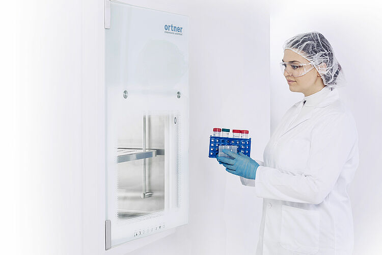 A woman in protective clothing stands in front of a closed Comfort S6 material pass. She holds laboratory equipment in both hands.