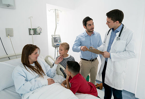 In einem Krankenhausbett liegt eine jung Frau. Neben dem Bett stehen ein Mädchen und ein Junge. Dahinter steht ein Mann, der gerade mit dem Arzt spricht.