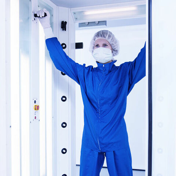 A woman stands in a chamber. There is a closed glass door on the back wall. The woman is dressed in a blue protective suit and wears a protective hood, cap and gloves in white. She has both her arms raised above her head and is holding on with her hands to black rods coming out of the side walls to her left and right. On the left edge of the chamber go from top to bottom evenly distributed several small black circles, from which comes a small white rod.