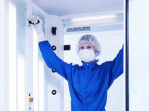  A woman stands in a chamber. There is a closed glass door on the back wall. The woman is dressed in a blue protective suit and wears a protective hood, cap and gloves in white. She has both her arms raised above her head and is holding on with her hands to black rods coming out of the side walls to her left and right. On the left edge of the chamber go from top to bottom evenly distributed several small black circles, from which comes a small white rod.