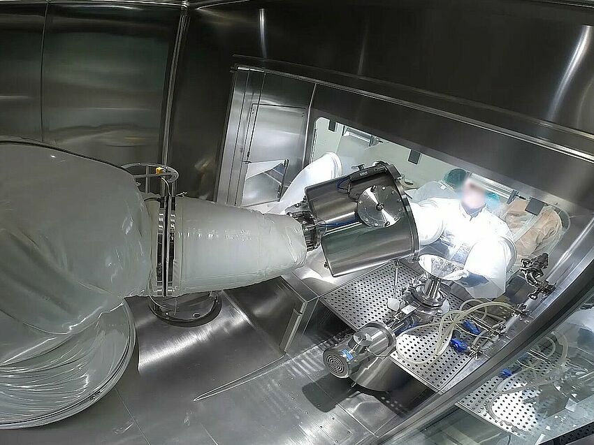 Zoom auf den Roboter im Innenraum des Isolators. Hinter der Glasscheibe sind außerhalb des Isolators Personen zu sehen, die durch Handschuhe in der Frontscheibe in das Innere des Isolators greifen.