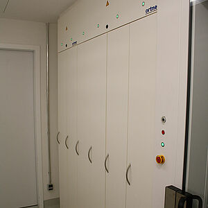  A large built-in cabinet, which consists of six narrow closed doors. Above is attached a closed box. On the right side is attached a box, also closed, which has a yellow-red, a green, a red and a black button halfway up. The green one lights up, and next to it is a closed glass door with a black handle.