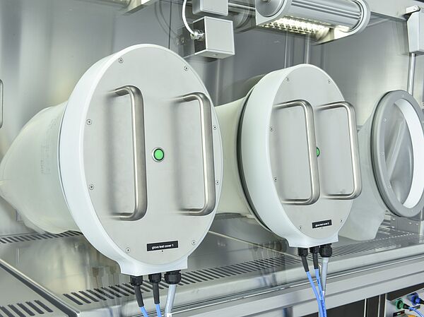 Two white discs are mounted in the closed front panel of an insulator. On the outside of each disc are two handles, and on the inside of each is a white glove that protrudes into the insulator's inner chamber. To the right of the two white discs is a circle, from which another glove also protrudes into the insulator's inner chamber.
