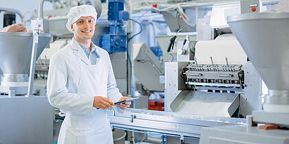 In einer Produktionshalle steht vor einer großen Maschine steht ein junger Mann in heller Kleidung und Schürze. Er hält ein Tablet in den Händen.