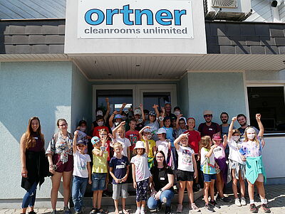 Am Haupteingang der Firma Ortner stehen Josef Ornter, drei Männer und drei Frauen umringt von Kindern, von denen ein paar ein Windspiel in Form eines Herzens in die Luft halten.