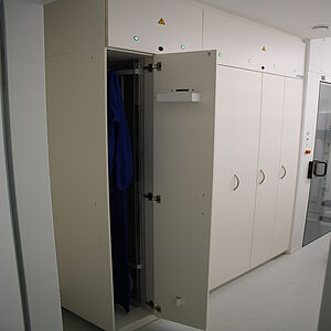  Along the left wall is a floor-to-ceiling cabinet with narrow cabinet doors. The leftmost closet door is open. A blue garment hangs inside. The upper part of the cabinet consists of two separate closed cabinet doors that can be opened upwards.