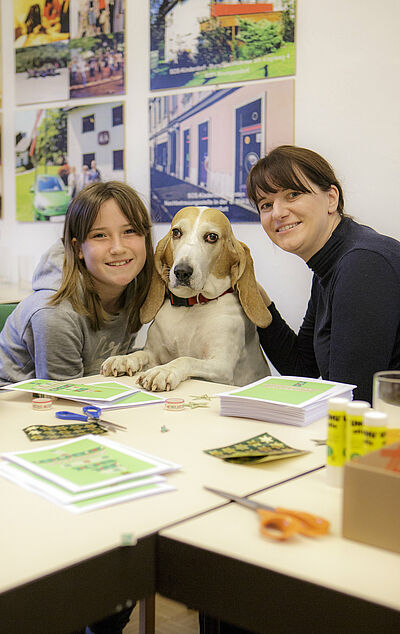 Stefanie Rud is sitting at a table with a dog. In front of them are brochures, scissors, glue and stickers.