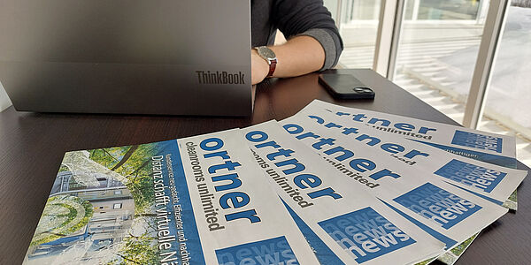 Am Tisch liegen aufgefächert fünf Ortner Zeitschriften mit dem Titel 'Ortner News'. Dahinter steht ein aufgeklappter Laptop an dem jemand arbeitet.