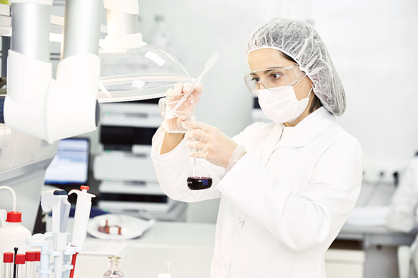Eine Frau in Labor-Kleidung füllt gerade Flüssigkeit von einem Laborbecher in einen Erlenmeyerkolben.