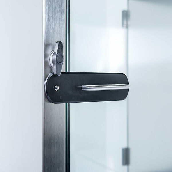 You can see the cutout of a door. A glass door is built into the silver door frame. On this glass door there is an elongated elevation with a silver thin handle. The elevation goes up to the door frame. Above this elevation is an elongated knob that goes from top to bottom and extends above the elevation.