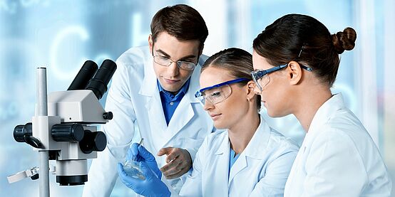 An einem Tisch sitzen zwei Frauen mit weißen Mänteln und Schutzbrillen. Die rechte Frau hält eine Petrischale in den Händen. Rechts von ihr steht ein Mann, der auf die Petrischale schaut.