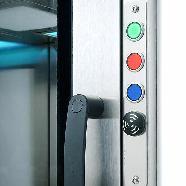  You can see the cutout of a small box with a closed door and large glass pane. A black handle is attached to the frame. To the right of it, from top to bottom, is a green, red, blue and black button. Through the glass pane you can see that the box is empty.