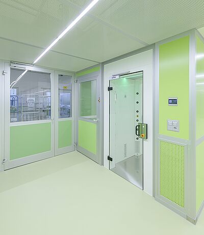 In der rechten hinteren Hälfte eines grünen Raums ist eine Personenluftdusche eingebaut. Zu sehen ist eine große geschlossene Türe aus Glas.