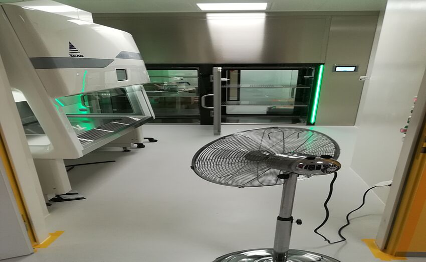 On the left is a workbench and next to it is a fan. At the end of the room are two airlocks whose doors are open. At the other end of each airlock is a closed glass door.