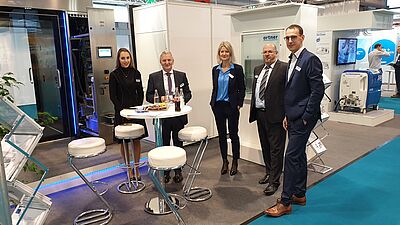 An Ortner employee, Josef Ortner, Lucia Matejcek, Klaus Gretter and Wolfram Kofler are standing at an exhibition stand. In the background is a Comfort S6 material pass-through.