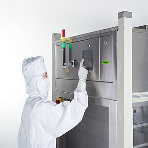  A person wearing a protective suit, hood, gloves and a white face mask is standing in front of a box that is slightly higher than the person. The lower part has a large glass pane. In the upper part, a rod protrudes upwards from the left side, which is black up to the half. The upper part is green from bottom to top, then yellow, then red. Next to it is a small screen. The person taps the screen with his right index finger.