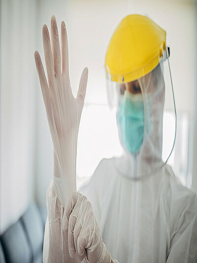 Eine Person mit weißer Schutzkleidung, Mundschutz und Helm mit durchsichtigem Schutzschild zieht sich gerade einen Schutzhandschuh an.