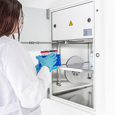 Vor einer geöffneten Materialdurchreiche steht eine Frau mit weißem Mantel und blauen Schutzhandschuhen. Sie hält eine kleine blau Box in den Händen, in dem Laborröllchen stehen.