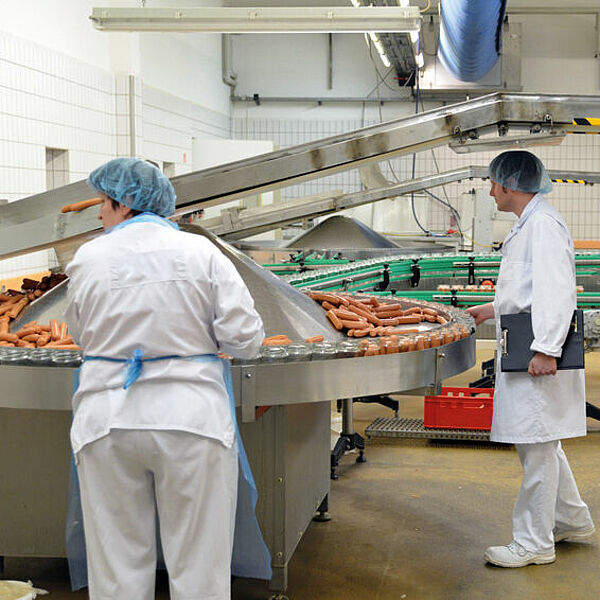  From the upper right corner of the picture, a conveyor belt goes to the left to the center. Below that is a conveyor belt that comes from the back left, curves eight and goes back to the back right. On it lie many small sausages. In front of it stands a woman in white clothes with an apron and a protective hood in blue. On the right side of the picture is a man with shoes, pants and coat in white, a protective hood in blue and a clipboard in his left hand.