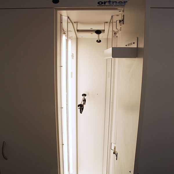 The inside of a locker. On the ceiling of the cabinet compartment is a handle from which hangs a hanger. On the left wall of the cabinet is a glass plate, behind which are switched on light tubes.