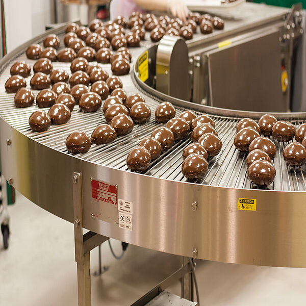 On a conveyor belt coming from the top right, making a turn to the bottom right, are many small cakes completely covered in chocolate.