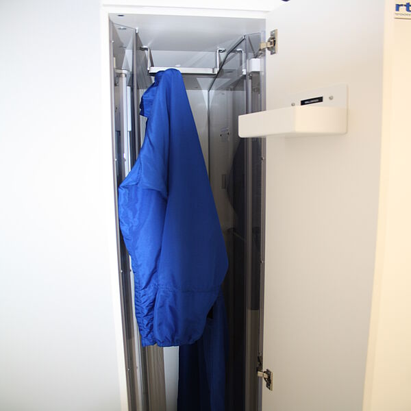  An open narrow closet. In the closet compartment hangs on a handle coming from the ceiling a blue jacket. On the left and right of the cabinet walls are glass panels.