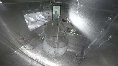 Der Innenraum eines Isolators, in der ein Roboter ist, der gerade eine Flüssigkeit versprüht. An der linken Wand ist eine Einbuchtung zu sehen, an deren Ende eine Glasscheibe ist, an der vier Handschuhe ins Innere ragen. Daneben steht hinter einer Glastür eine Person, die den Roboter beobachtet.