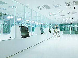 A large white room with 14 separated high glass panes. Every fourth pane of glass has a box built into the lower part in landscape format. At the box at the very front, the door, which is completely made of glass, is open. The door has a black handle. Behind the glass panes you can see another large white room.
