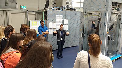 In the production hall in Möllbrücke, Katharina Ortner and Michael Ranacher are standing in front of a personnel lock and a group of young women. Katharina Ortner is explaining something.