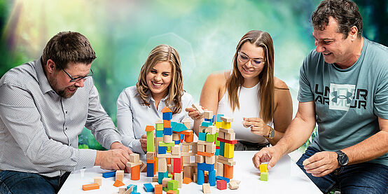 Against a green background, four people sit side by side at a white table. Towers are built out of colourful building blocks on the table. On the far left is a man in a black-and-white patterned shirt and glasses. Next to it sits a blonde woman in a black and white striped blouse, next to it sits a brunette woman with glasses and a sleeveless white shirt. Next to it is a man with black hair and a blue shirt. Everyone laughs.