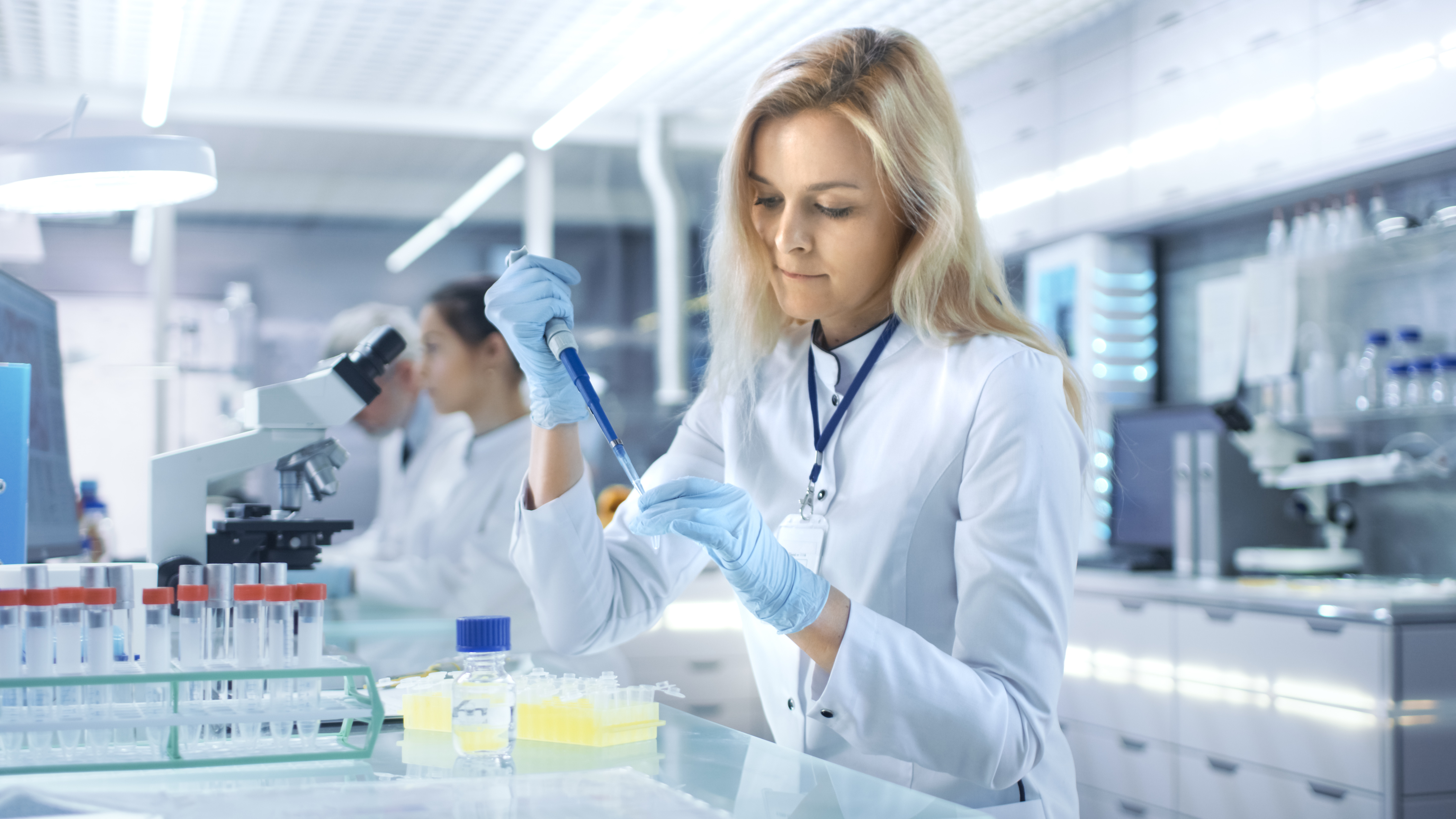 Eine blonde Frau in weißer Kleidung hält in der rechten Pipette, mit der sie gerade etwas in ein kleines Gefäß in ihrer linken Hand füllt. Ein Stück weiter rechts ist ein Mikroskop und sind eine Frau und ein Mann zu sehen.